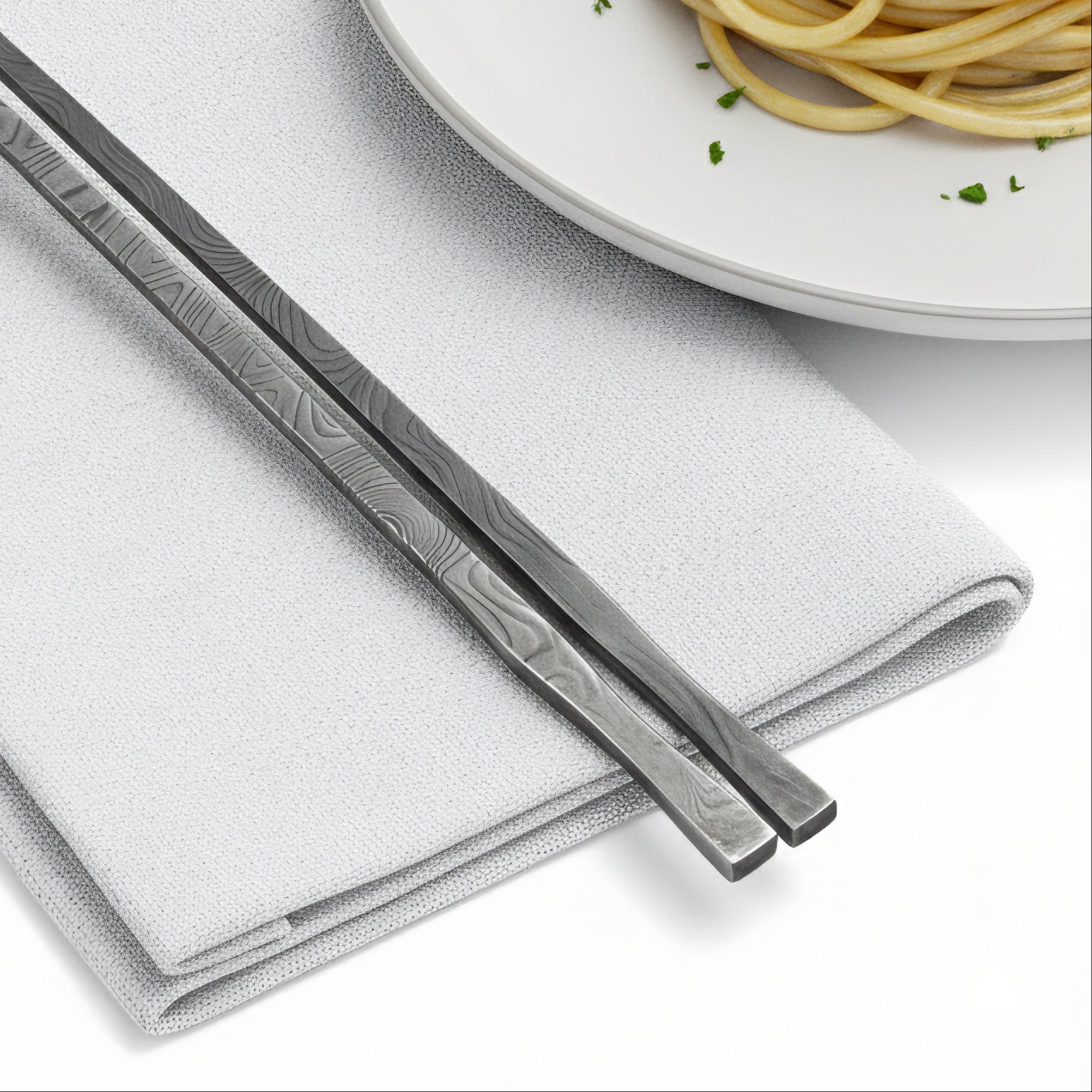 Decorative metal chopsticks on a folded white napkin with a plate of pasta in the background.