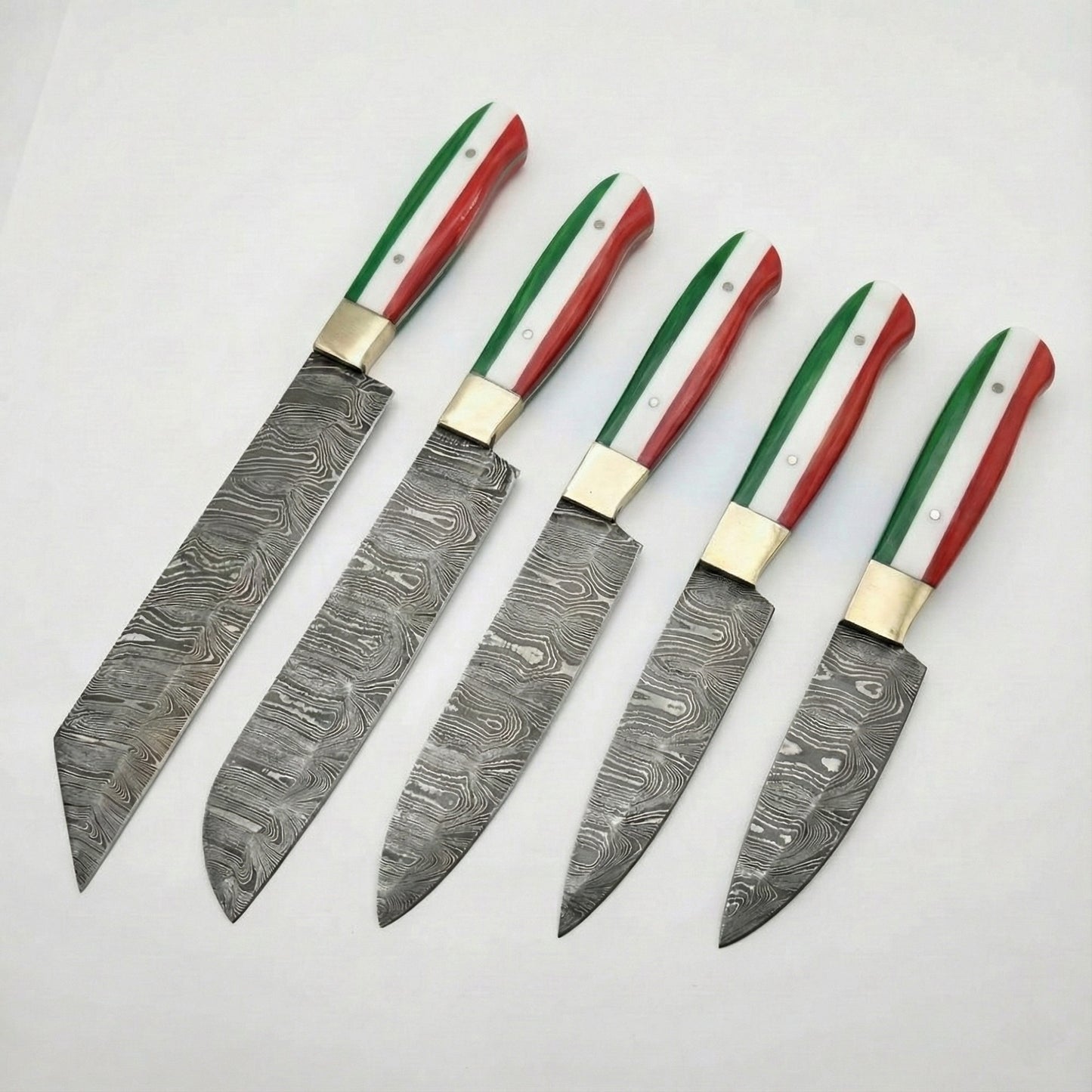 A set of five custom kitchen knives arranged diagonally on a white background. Each knife features a Damascus steel blade with a swirling, wavy pattern and a brass bolster. The handles are uniform across the set, designed with vertical stripes of green, white, and red to represent the Italian flag. The blades vary in shape and size, including a large cleaver-style blade, a chef's knife, and smaller utility or paring knives.