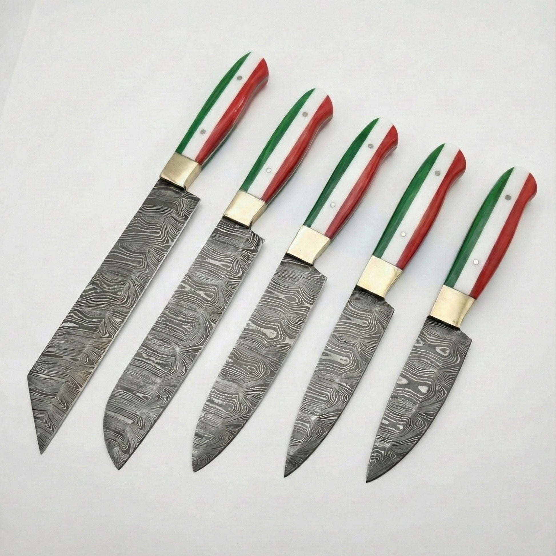 A set of five custom kitchen knives arranged diagonally on a white background. Each knife features a Damascus steel blade with a swirling, wavy pattern and a brass bolster. The handles are uniform across the set, designed with vertical stripes of green, white, and red to represent the Italian flag. The blades vary in shape and size, including a large cleaver-style blade, a chef's knife, and smaller utility or paring knives.