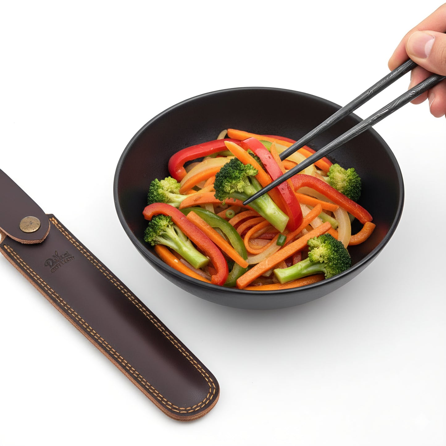 Black bowl with stir-fried vegetables and a pair of chopsticks, next to a leather case on a white background.