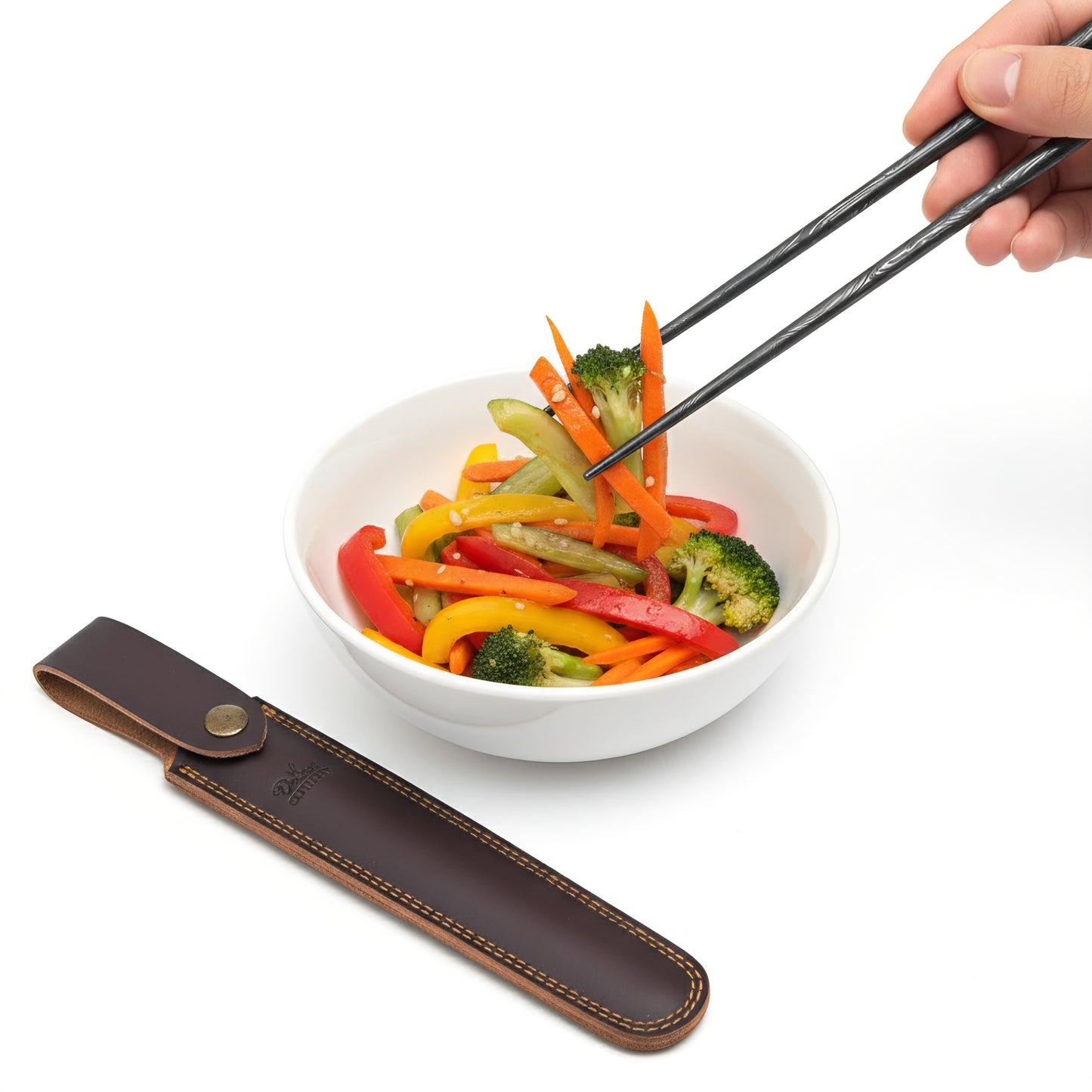 Bowl of colorful vegetables with a pair of black chopsticks held by a hand, next to a brown leather chopstick holder on a white background.