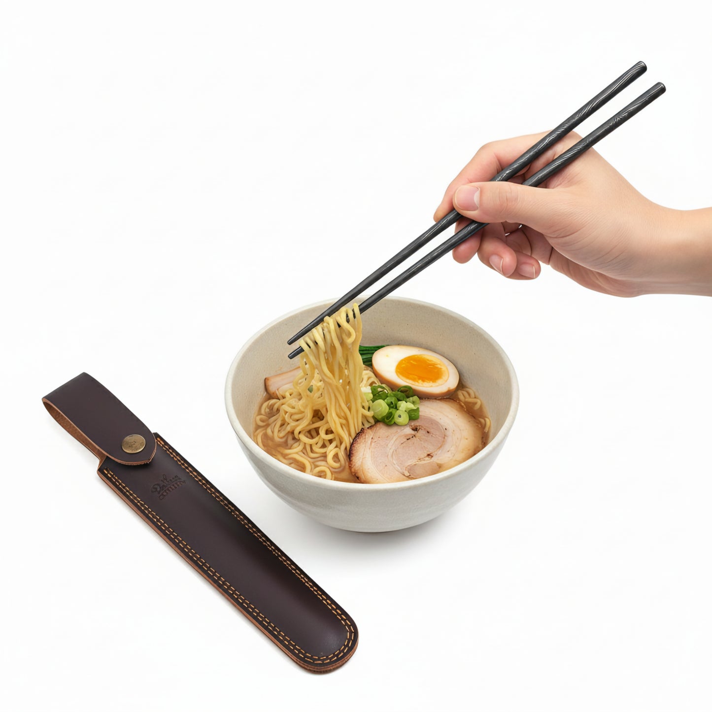 Hand holding chopsticks over a bowl of ramen with a leather case on a white background