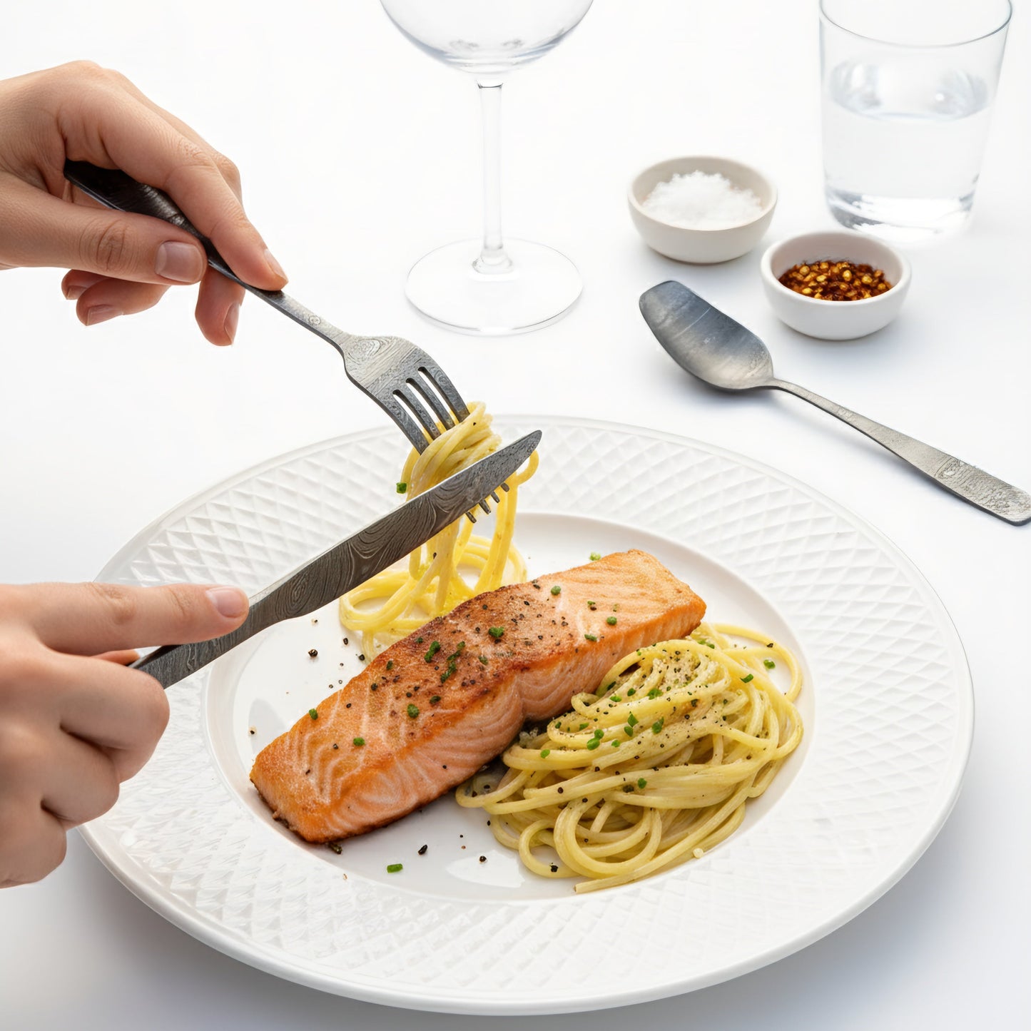 Person cutting into a plate of salmon and pasta with cutlery on a white Damascus cutlery set table.