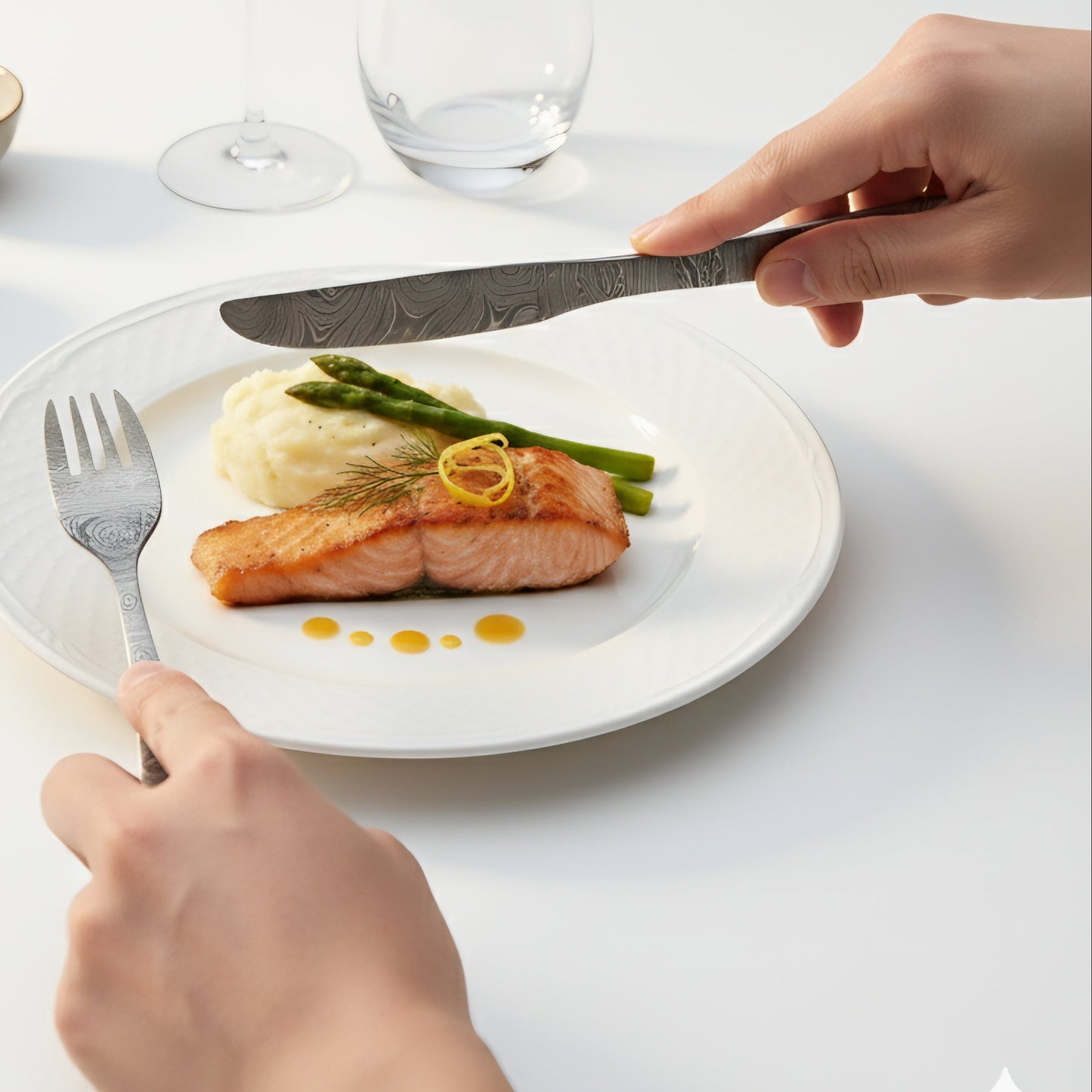 Person cutting into a plate of salmon with asparagus and mashed potatoes on a white table. Damascus cutlery set